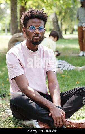 Portrait of African young man in eyeglasses looking at camera while sitting on the grass in the park Stock Photo