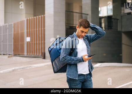 Front view of confused handsome male courier with thermo backpack using navigation app on phone standing in city street. Stock Photo