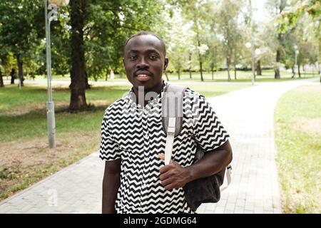 Portrait of African student with backpack behind his back smiling at camera while walking through the park Stock Photo