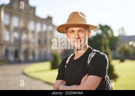 Travel and old city explore concept. Cheerful man traveler in hat and backpack in European city center background. Portrait of young adventurous traveler or wanderlust on vacation. High quality photo Stock Photo