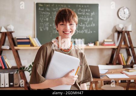 Portrait of happy young teacher with books smiling at camera while standing in empty classroom at school Stock Photo