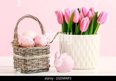 Pink Easter decorations: tulip flowers, basket with painted eggs and dotted rabbit figure on white wooden table. Stock Photo