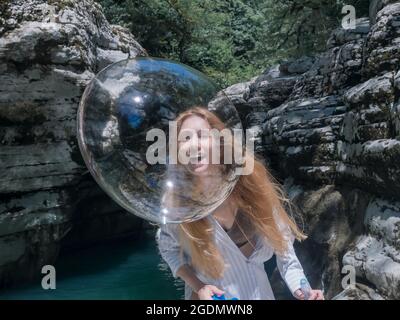 Laughing girl catches up with shiny soap bubble looking through it against the backdrop of canyons and water Stock Photo