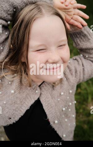 Top view of adorable little girl in knitted jacket squinting and smiling while resting on grassy ground in countryside Stock Photo