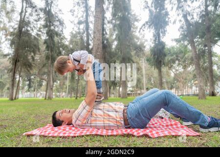Man lying on a checkered picnic tablecloth in a city park as he lifts his son with his arms. Stock Photo