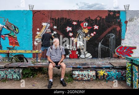 Elderly man resting on bench in front of Young artist painting new art on wall in Mauerpark, Prenzlauer Berg, Berlin Stock Photo