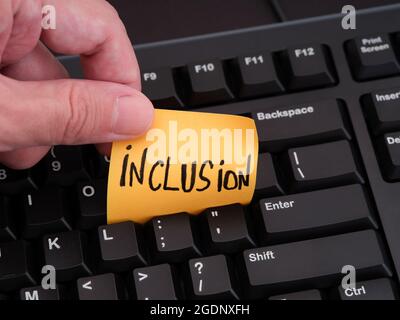 A person holding a piece of paper with the word Inclusion on it between the keys of a black computer keyboard. Close up. Stock Photo