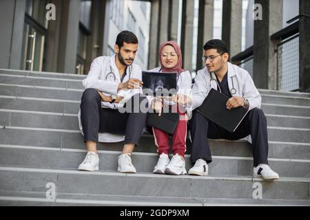 Medical team of three diverse Arabian doctors surgeons, sitting on the stairs outdoors of modern hospital and looking at skull X-ray, discussing ways of treatment and operation Stock Photo