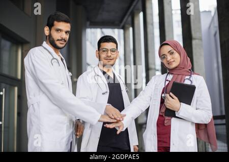 Motivated medical team of three Arab doctors, showing their unity and collaboration. Two male handsome doctors and their pretty female colleague in hijab, stacking hands, standing outside clinic Stock Photo