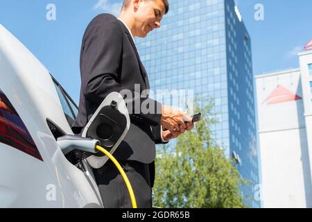 Man in a black suit, standing near an electric car that is charging and making time adjustments on a smartphone Stock Photo