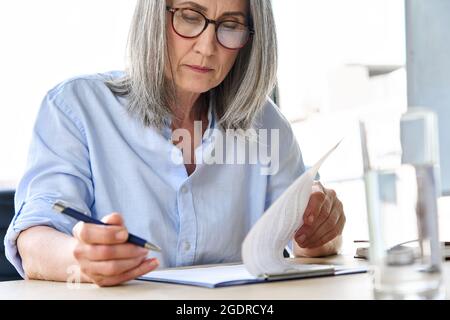 Focused older businesswoman in glasses reading partnership contract at table. Stock Photo