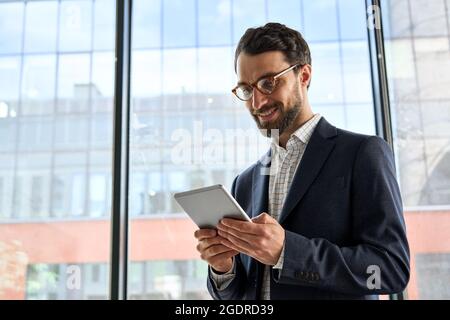 Smiling mature bearded businessman holding tablet device standing in office. Stock Photo