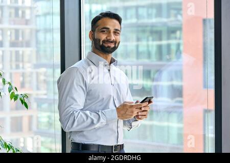 Smiling indian businessman holding phone standing in office at panoramic window. Stock Photo