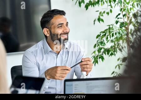 Smiling male Indian ceo listening colleague holding pen in hands at table. Stock Photo