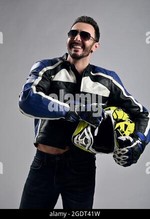 Laughing bearded man biker motocross race winner in motorcycle leather jacket and gloves stands holding his helmet Stock Photo