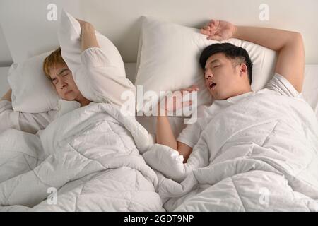 Asian man snoring loudly while his wife is covering ears with the pillow Stock Photo