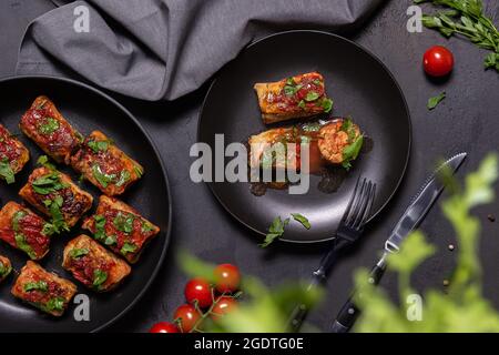 Flat lay with stuffed cabbage rolls in tomato sauce and chopped parsley in black crockery, linen napkin and some ingredients on side. Unfocused parsle Stock Photo