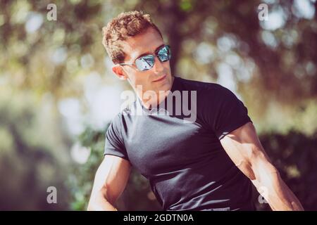 Charming young man with sunglasses and black t-shirt outdoors in a park with intense sunshine. Stock Photo