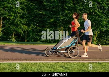running couple with child in jogging stroller in public park at sunrise. Morning run for a whole family Stock Photo