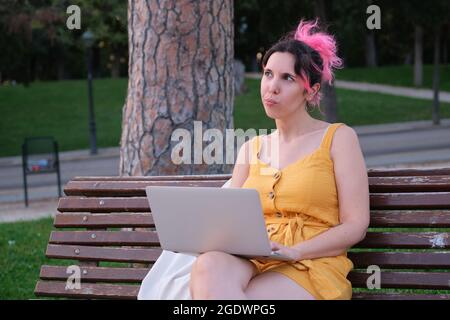 Young caucasian woman with pink hair thinking while working on a laptop. Stock Photo