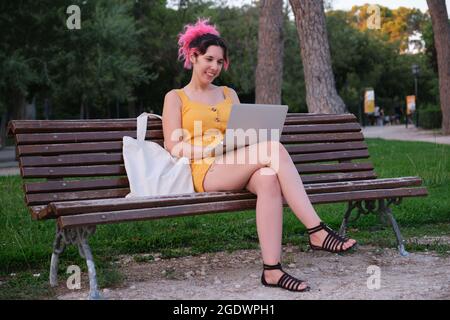 Young caucasian woman with pink hair working on a laptop in a park bench. Stock Photo