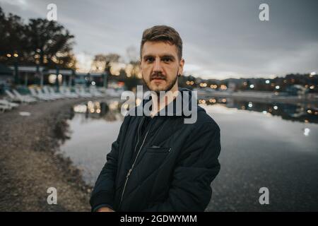 A shallow focus shot of a Caucasian guy wearing a coat looking at the camera standing on a shore Stock Photo