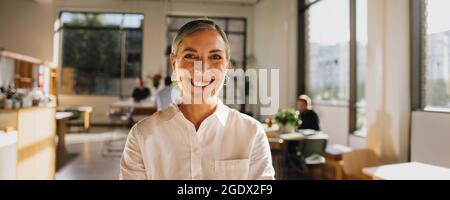 Positive female entrepreneur standing and smiling in co-working space with people in background. Happy senior manager standing in office. Stock Photo