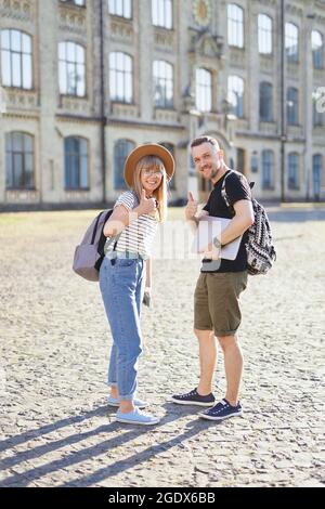 International successful students couple smiling showing thumbs up sign at the camera. Two attractive students, male and female, with backpacks showing thumbs up gesture at university campus Stock Photo