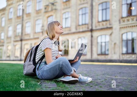 Browsing internet, blogging or social network usage concept. Cute successful caucasian blonde female student at university campus sitting on green grass with backpack and laptop. High quality photo Stock Photo