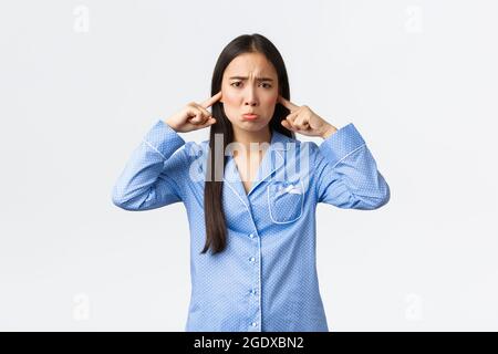 Timid and sad asian girl in blue pajamas pouting and whining, shut ears with fingers and sulking displeased, feeling discomfort from disturbing sound Stock Photo