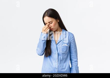 Distressed and exhausted asian girl feel bad, wearing pajamas cant sleep at night, rubbing closed eyes, having insomnia, standing unwell with headache Stock Photo