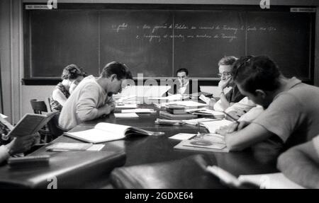 1950s, historical, some college students in a classroom sharing a large desk studying French literature, with the male college professor sitting reading infront of the blackboard, USA. Stock Photo