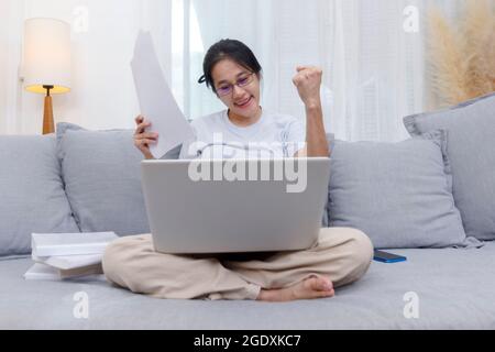 Asian woman celebrating online win success looking laptop at home. Work from home business women sitting on sofa in living room working with laptop on Stock Photo