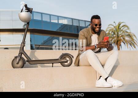 Business african american man having fun using mobile phone with electric scooter outdoor in the city - Focus on face Stock Photo