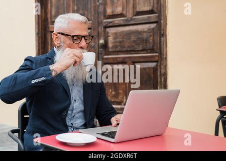 Senior hipster business man using laptop computer while drinking coffee outdoors at cafe bar - Joyful elderly lifestyle and technology concept - Focus Stock Photo
