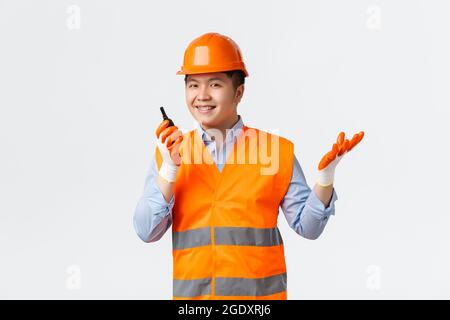 Building sector and industrial workers concept. Smiling confident asian chief engineer, construction manager give orders via walkie-talkie, looking Stock Photo