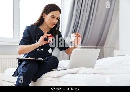 The brunette woman doctor sitting on bed in front of a laptop and showing two jars of pills to the laptop camera on the background of the room Stock Photo