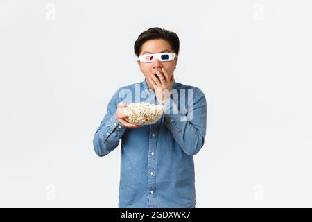 Leisure, lifestyle and people concept. Astonished asian guy in 3d glasses munching popcorn excited looking at TV, watching movie on wide screen Stock Photo