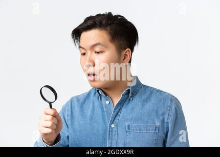 Close-up of young asian guy looking curious and focused, searching for something, looking through magnifying glass down, open mouth intrigued Stock Photo