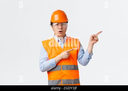 Building sector and industrial workers concept. Impressed and amazed asian construction manager, engineer in helmet and reflective clothing pointing Stock Photo