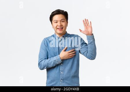 Smiling friendly-looking asian man being honest, raising arm and hold hand over heart while telling truth, making promise or swear something, standing Stock Photo