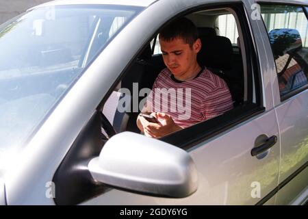 Successful caucasian young man sitting in car. 20s guy using mobile phone. Silver grey gray car. Attractive brunette men in vehicle. Buyer or auto dea Stock Photo