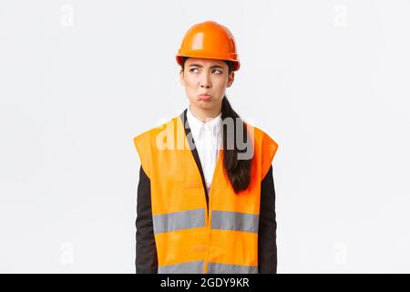 Gloomy and offended young asian female construction manager, industrial woman in safety helmet complaining or whining, pouting sad and looking upper Stock Photo