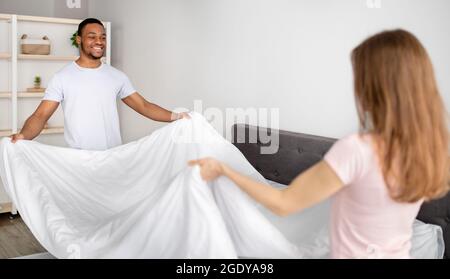 Positive multiethnic couple making bed in morning together at home Stock Photo
