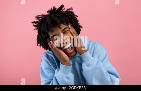 Funky African American teen opening mouth in shock, shouting WOW, holding his face, looking at camera on pink background Stock Photo