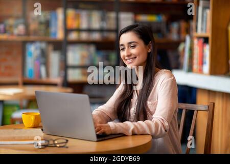 Remote learning and online work concept. Happy arab woman using laptop computer at cozy urban cafe, free space Stock Photo