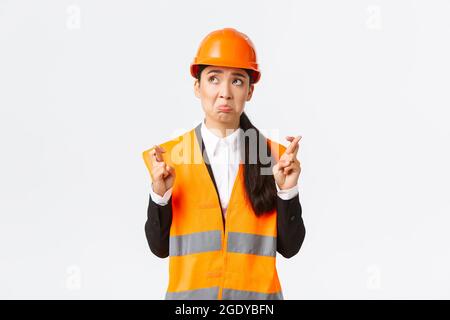 Sad nervous and unconfident asian female construction manager in safety helmet, pouting and look in panic upper left corner, making wish, cross Stock Photo