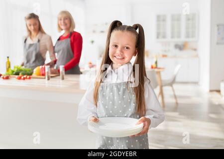Cute happy girl in apron holding clean plates while her grandma and mother preparing dinner at table in kitchen Stock Photo