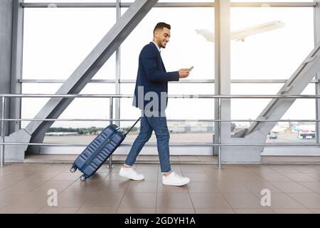 Handsome arab guy using smartphone while walking with suitcase at airport terminal Stock Photo