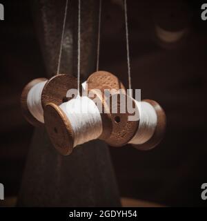 Selective focus foreground on hanging group of thread shabby bobbins with white cotton yarn on blurred dark brown background. Stock Photo